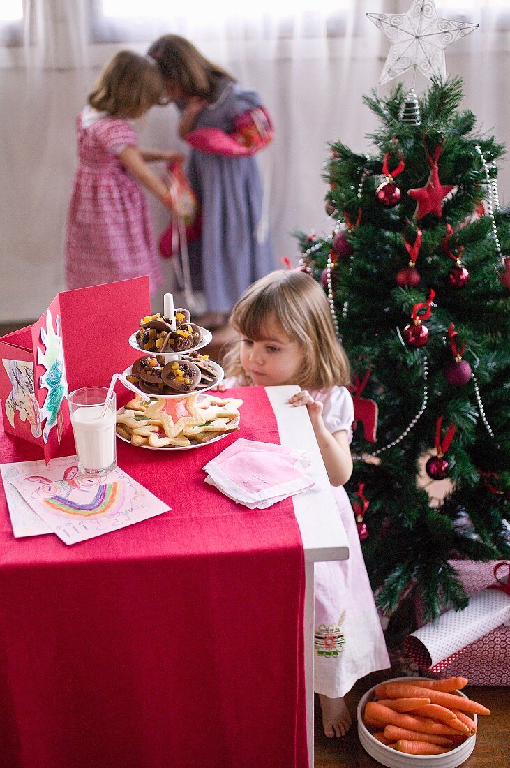 Kleines Mädchen vor geschmücktem Weihnachtsbaum auf eine Etagere voller Plätzchen blickend