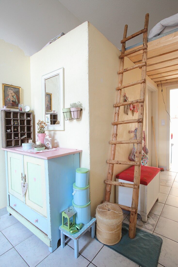 Rustic ladder leading to sleeping platform above doorway next to pastel chest of drawers
