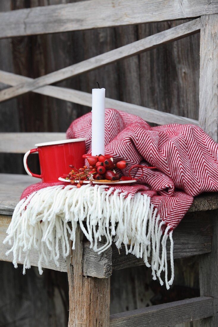 Candlestick decorated with rose hips next to mug arranged on fringed blanket and rustic wooden bench