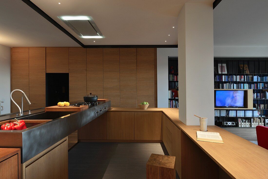 Offene Designerküche aus Holz mit Metallspüle und integriertem Pfeiler; Blick in Wohnbereich mit Regalwand