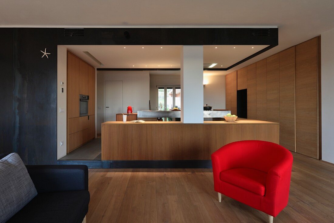 Moderne Küche mit schlichten Holzfronten in offenem Wohnbereich mit rotem Armlehnsessel und schwarzer Metall-Wandverkleidung