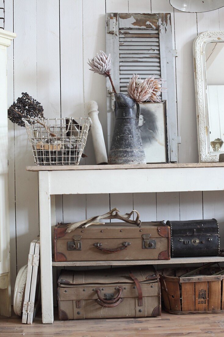 Old suitcases on shelves below console table decorated with vintage ornaments and jug of proteas