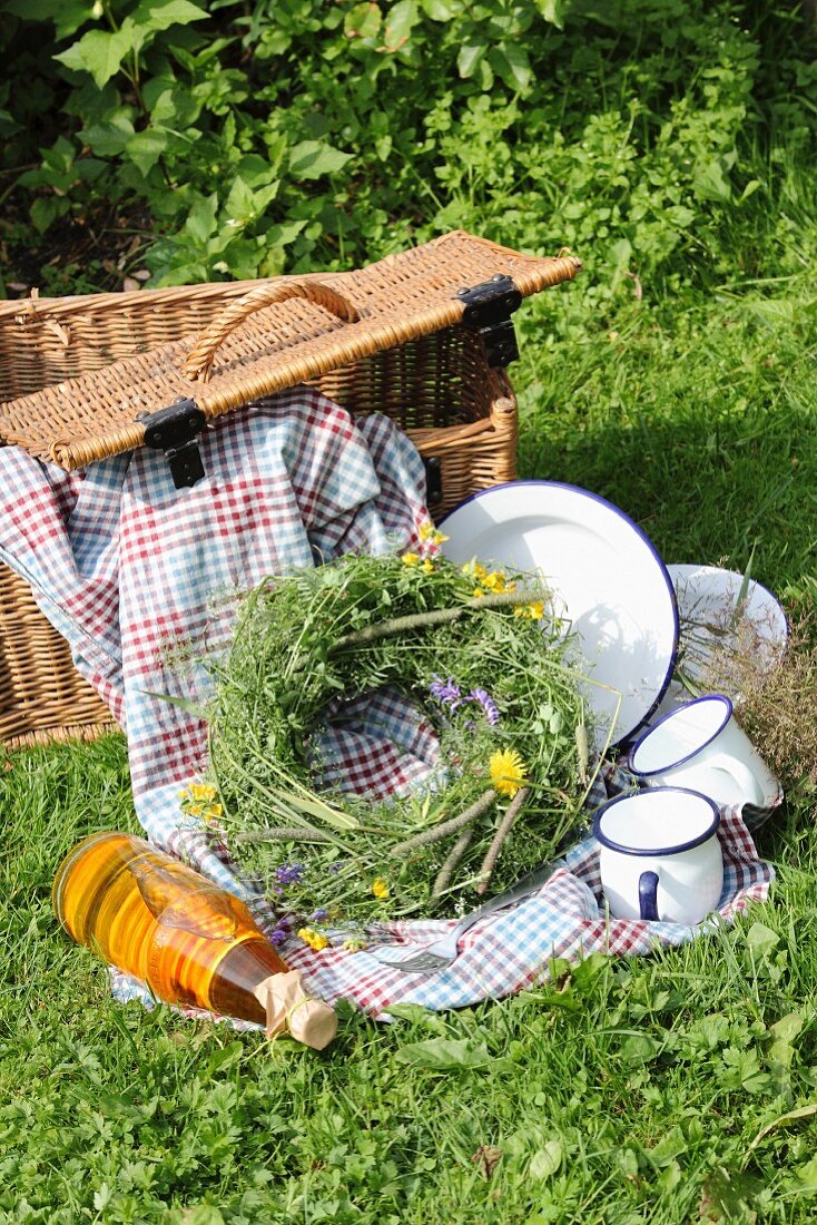 Picknickkorb mit Decke und Emaillegeschirr auf grüner Wiese mit Wiesenblumenkranz