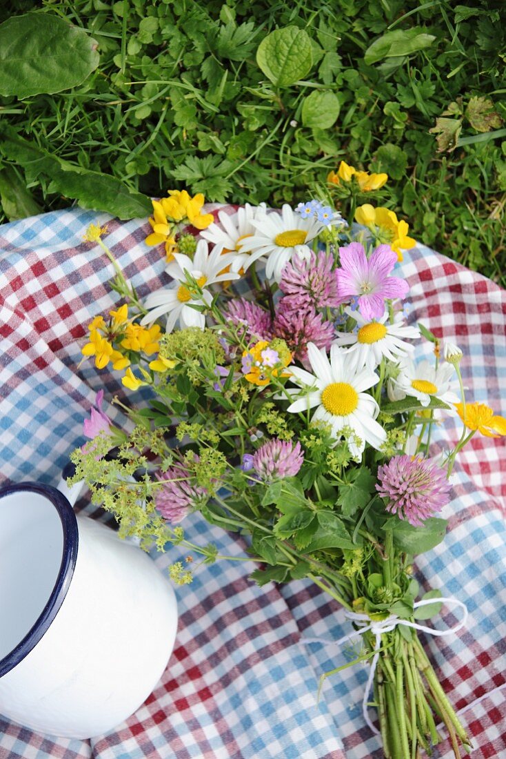 Karierte Picknickdecke mit buntem Wiesenblumenstrauss und weisser Emailletasse im Gras