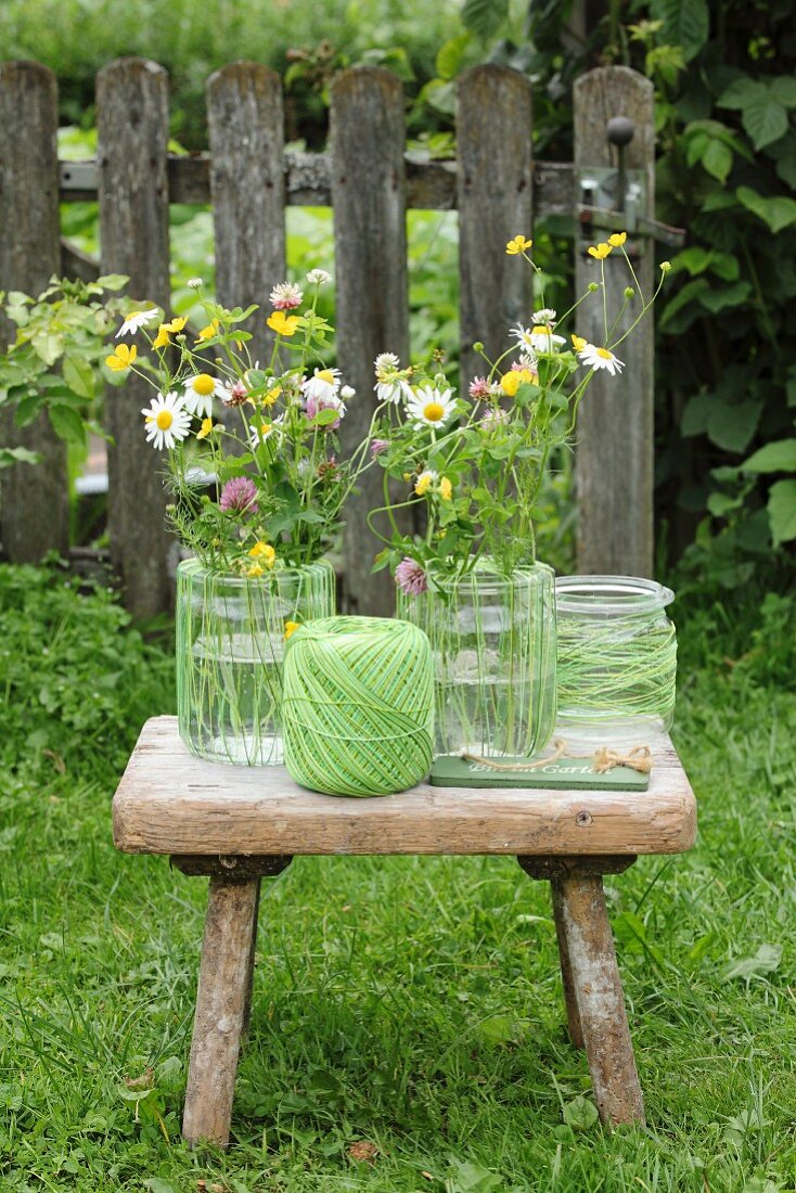 Still-life arrangement of glass vases, one wrapped in twine, and colourful wildflowers on vintage wooden stool
