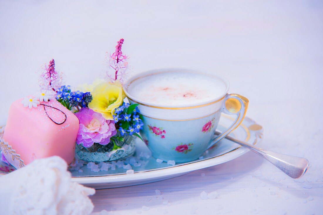 Eine Tasse Cappuccino, Blumengesteck und Petit Four