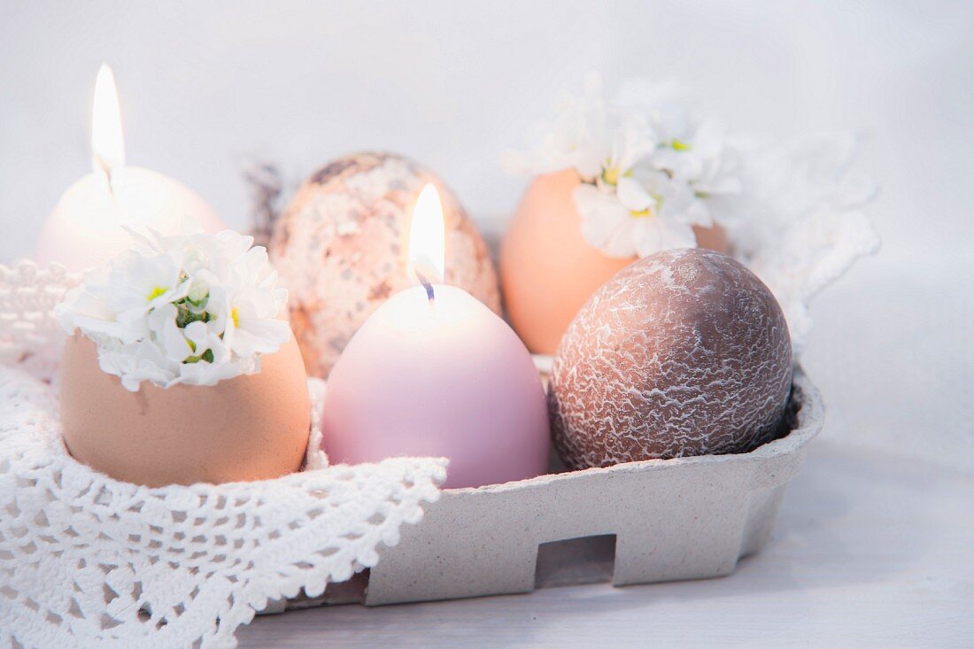 Eierkarton mit Eiern, brennenden Kerzen, weißen Blüten und Häkeldeckchen