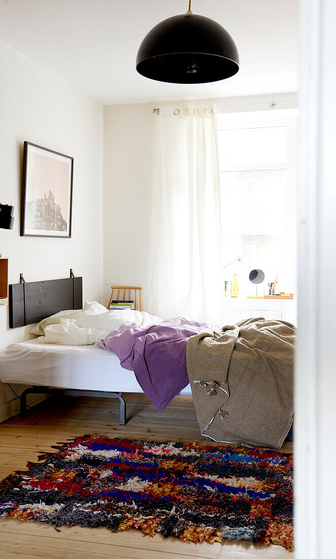 Bed with headboard made from stained wooden board and black leather straps and Moroccan rug on floor in bedroom