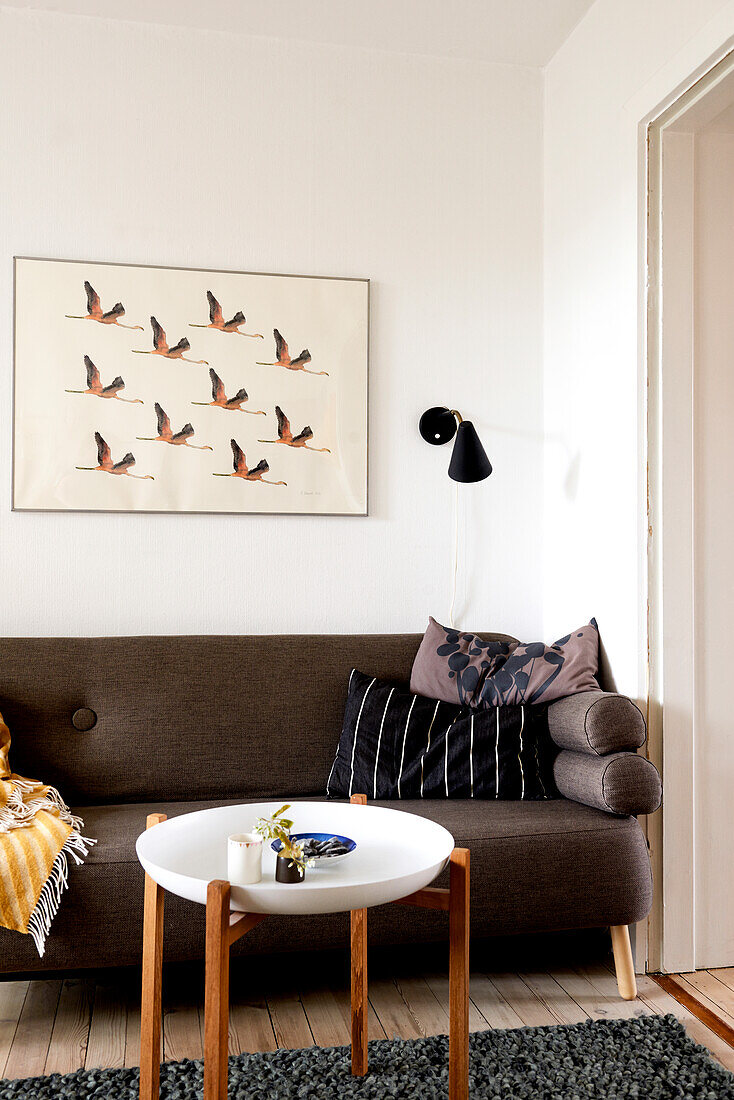 Side table, scatter cushions on brown sofa, picture of birds and sconce lamp