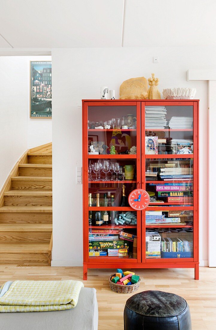 Games, glasses and magazines in red, glass-fronted cabinet next to foot of staircase