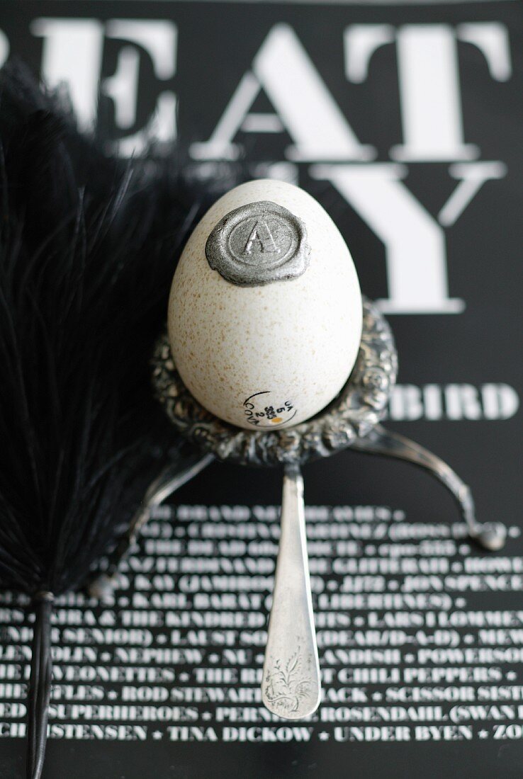 Blown egg with wax seal in silver egg cup on black and white lettered surface