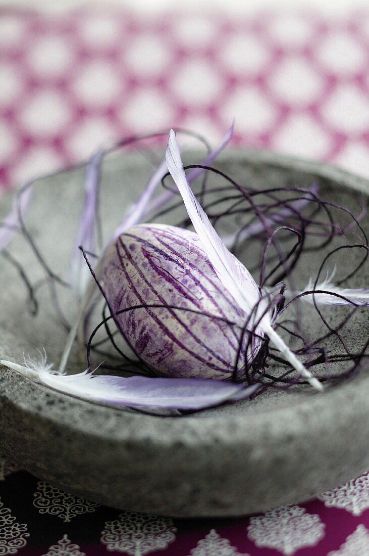 Easter egg painted purple, feathers and delicate cord in stone dish