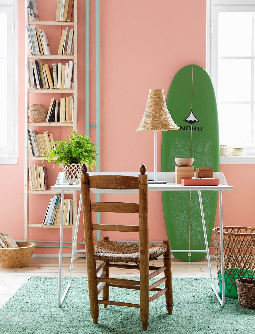 Einfaches Leiterregal für Bücher an apricotfarbener Wand neben angelehntem grünem Surfbrett, davor filigraner Schreibtischplatz mit rustikalem Holzstuhl