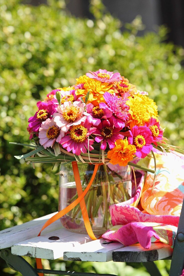 Farbenfroher Blumenstrauß mit Zinnias und Tagetes in Glasvase auf Vintage Gartenstuhl