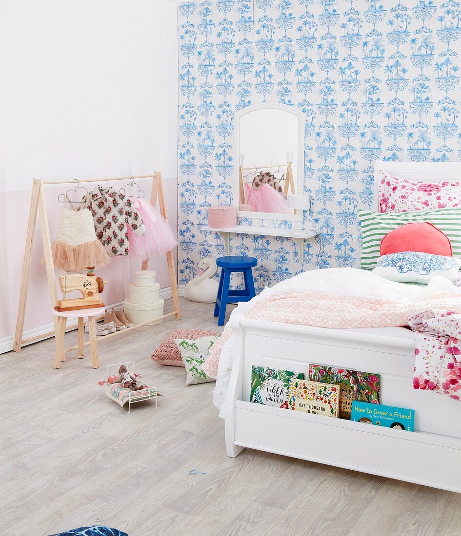 Weisses Bett mit Bücherfach vor blau-weisser Mustertapete und Kleiderständer mit Balettkleidung im Mädchenzimmer