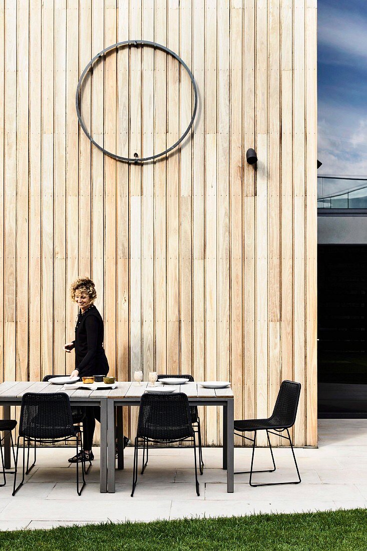 Langer Esstisch mit Stühlen vor Holzwand auf Terrasse, Frau im Hintergrund