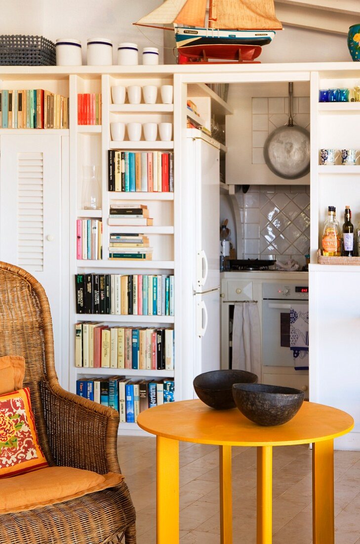 Comfortable wicker armchair, yellow side table, bookcase and kitchen area