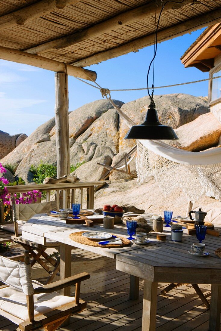 Esstisch und Stühle auf rustikaler Terrasse mit Hängematte neben Fels und mediterranem Flair