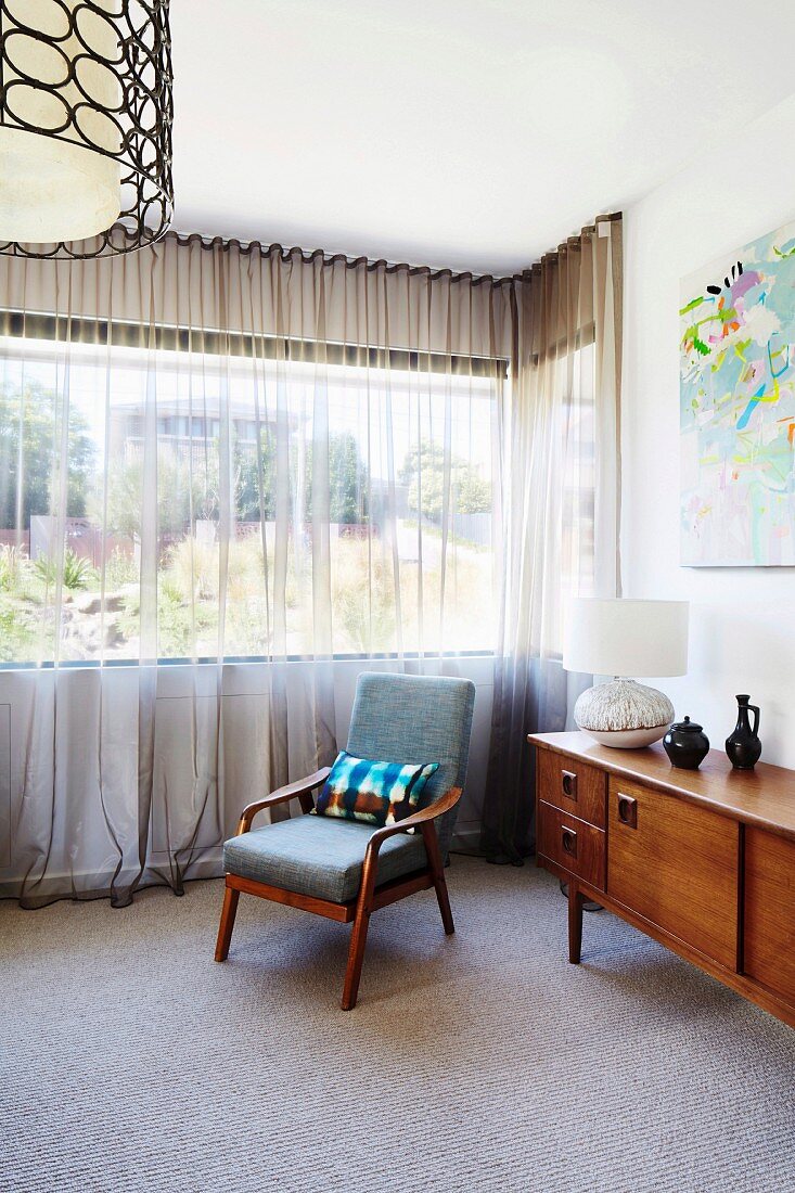 Gepolsterter Armlehnstuhl und Sideboard im Fifties Stil vor Fenster mit luftigem Vorhang