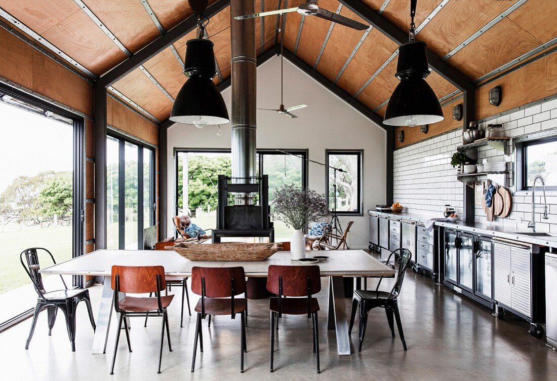 Open kitchen in industrial style, with concrete floor, underground tiles and wood-clad ceiling, in the foreground a long dining table with chairs