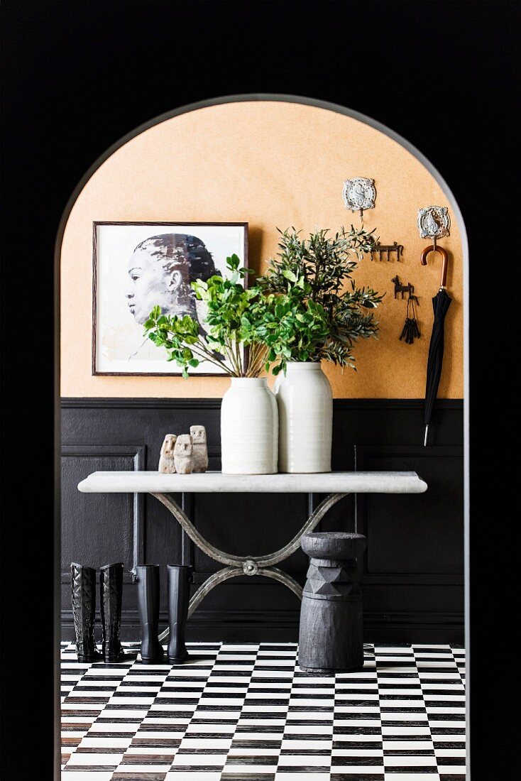 Ceramic vases with leaf sprigs and Sumba satutes on a console table, in front of which black boots on a black and white tiled floor