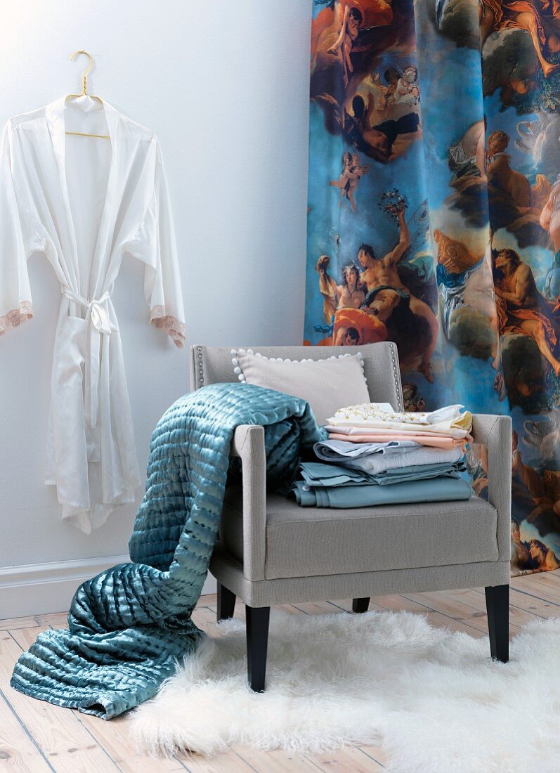 Grauer Polstersessel mit Bettwäsche und glänzender Tagesdecke vor Vorhang mit Engelsmotiven, an Wand Kimono im Ankleidezimmer