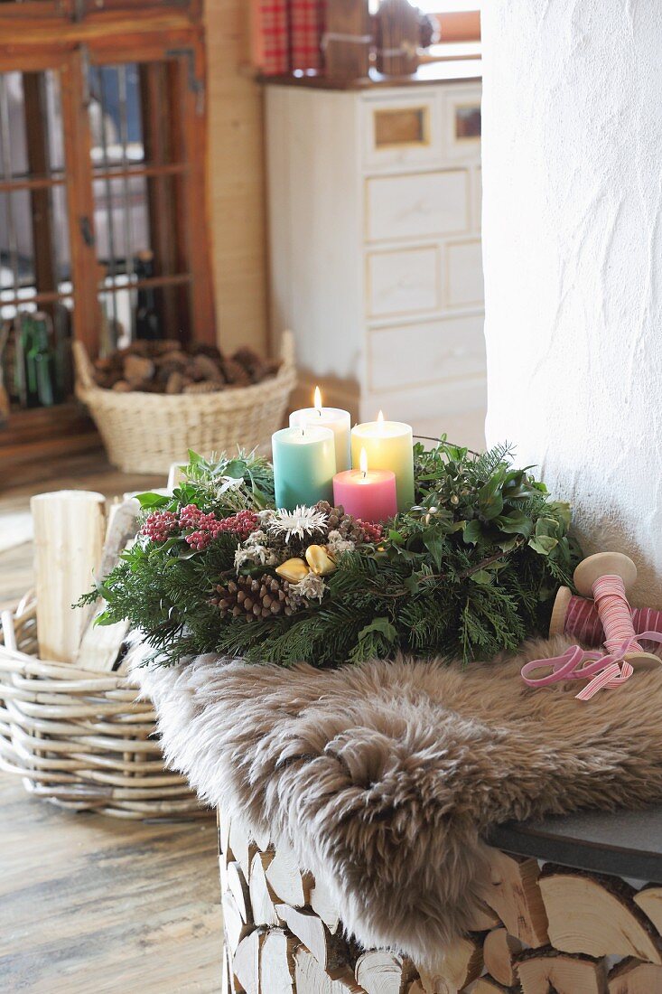 Advent wreath with four colourful candles on sheepskin rug on bench next to fireplace