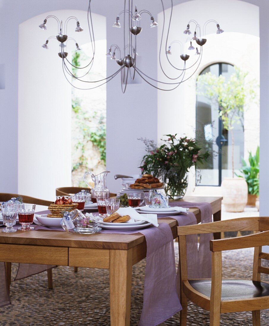 Gedeckter Eichentisch mit lilafarbenen Tischläufern unter Halogen-Kronleuchter im mediterranen Innenhof