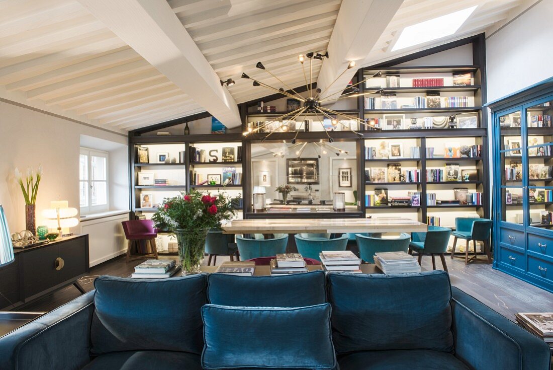 Blue sofa, long table, shell chairs and wall covered in shelves in open-plan interior