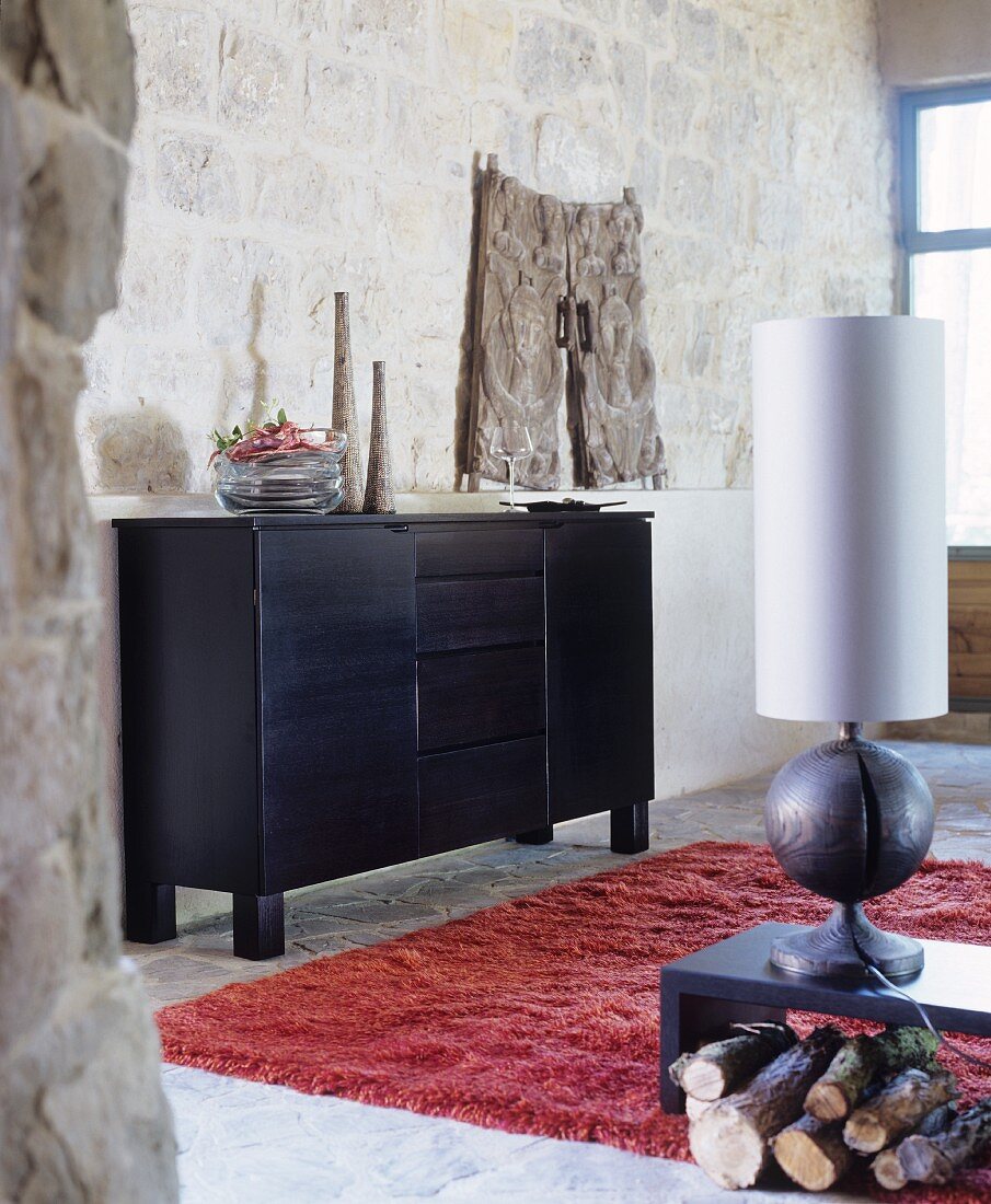 Black sideboard, table lamp with wooden base and red flokati rug in rustic living area