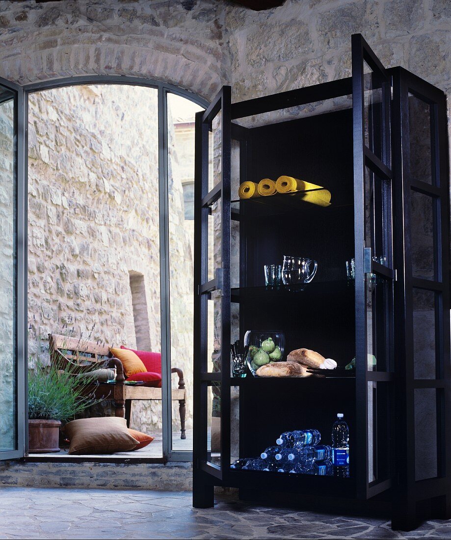 Bottles of water and bread in black, glass-fronted cabinet next to open French windows with view of stone wall and wooden bench
