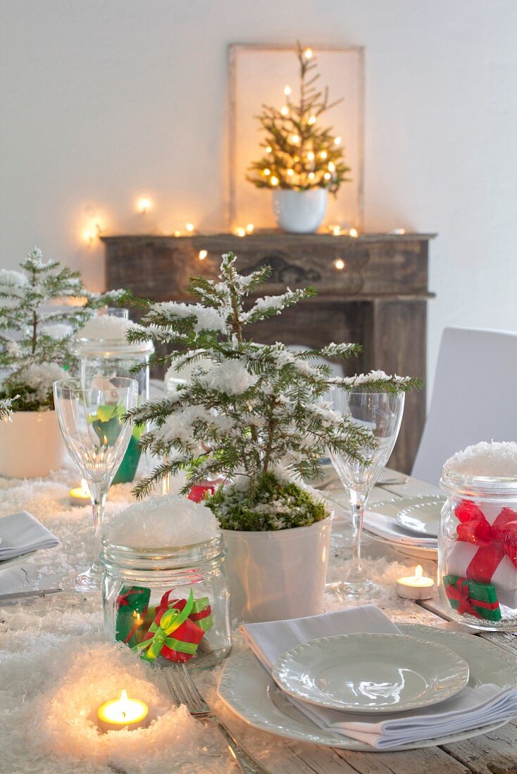 Weihnachtstisch mit Dekoschnee, Fichtenbäumchen, weißem Geschirr und brennenden Teelichtern