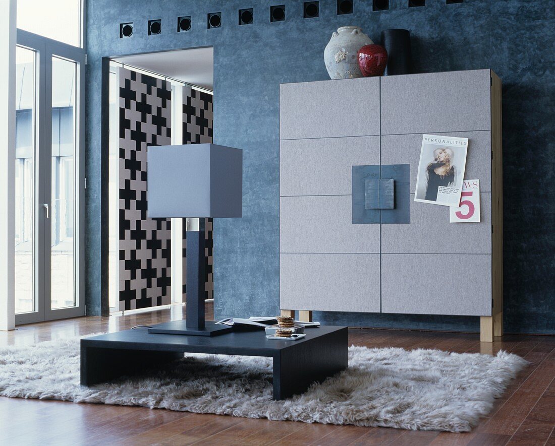 Grey cupboard with felt-covered front against blue-grey wall and table lamp on low table on flokati rug