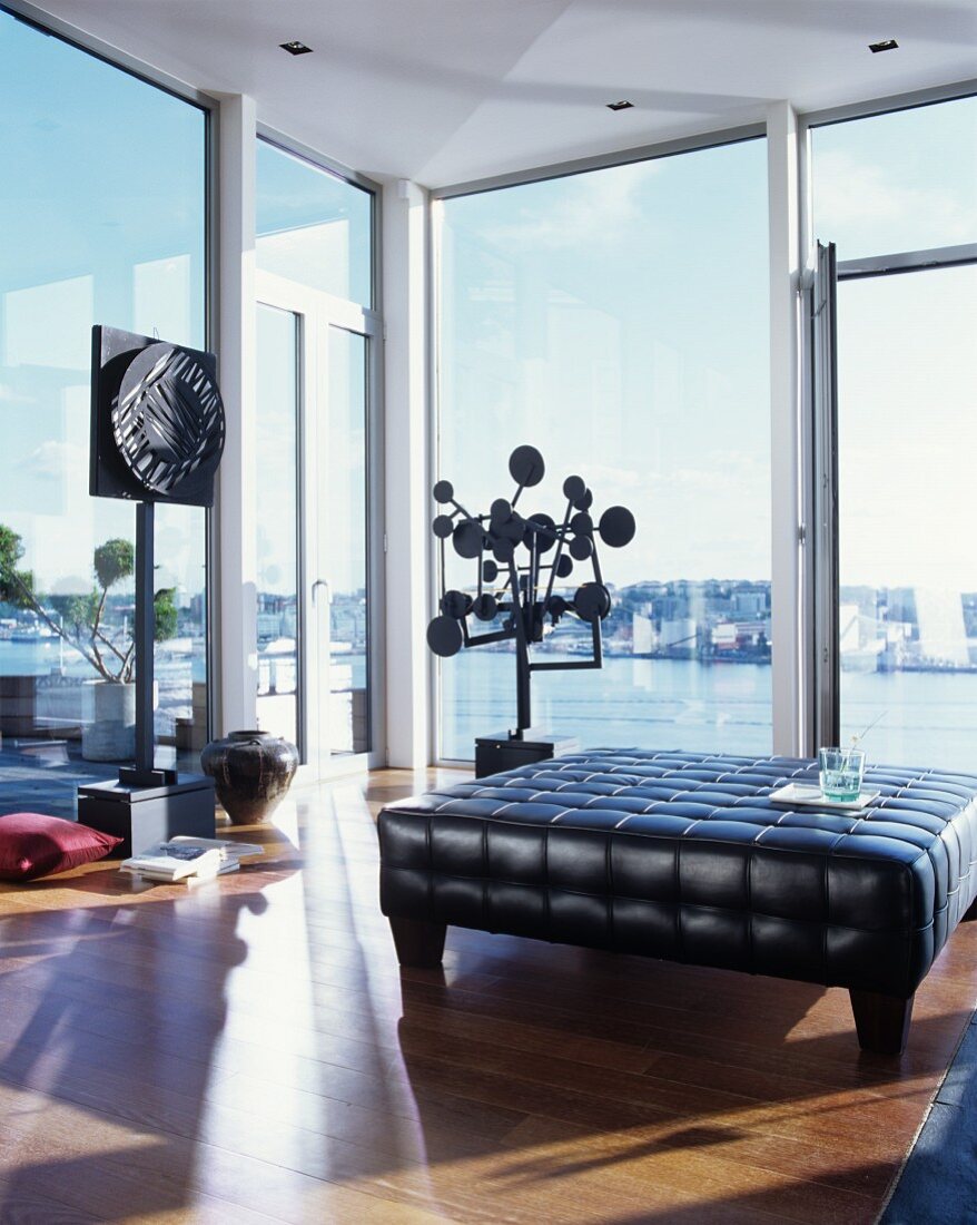 Schwarzer Leder-Polstertisch vor modernen Kunstobjekten und Glasfront mit Seeblick