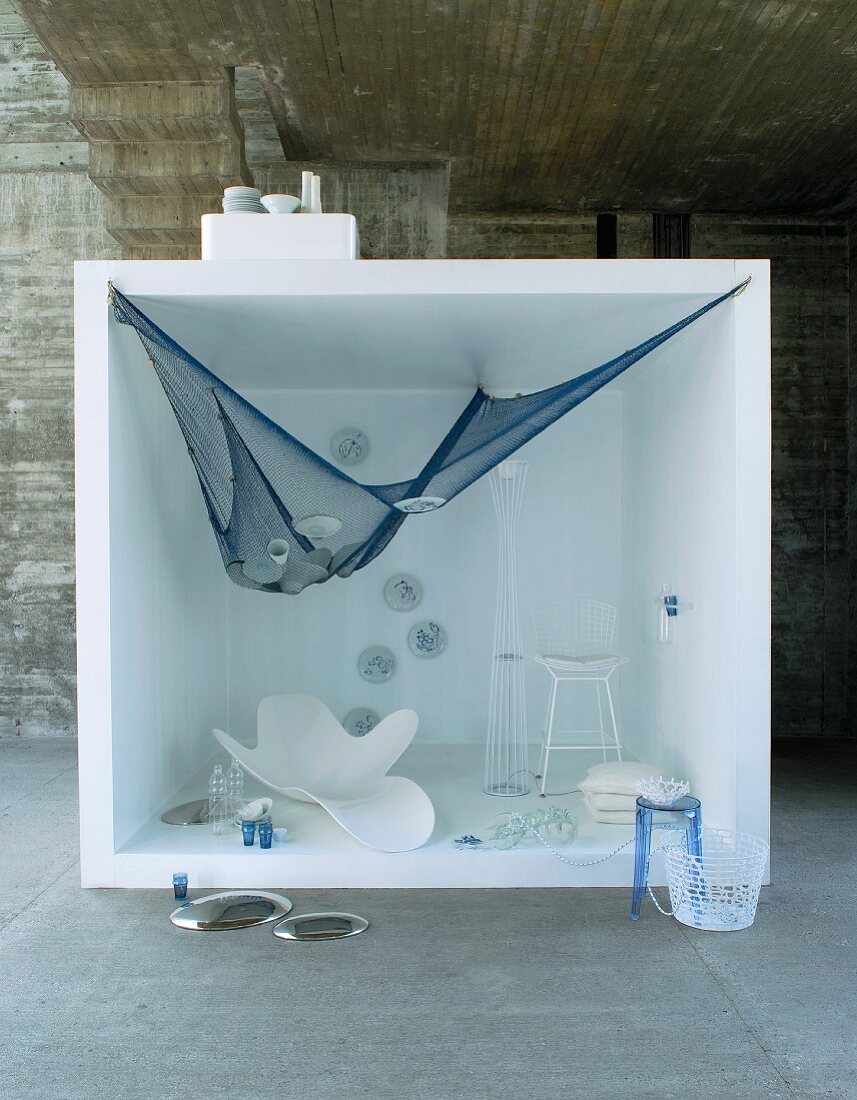 White furniture and blue fishing net hung up in white cube in front of concrete wall