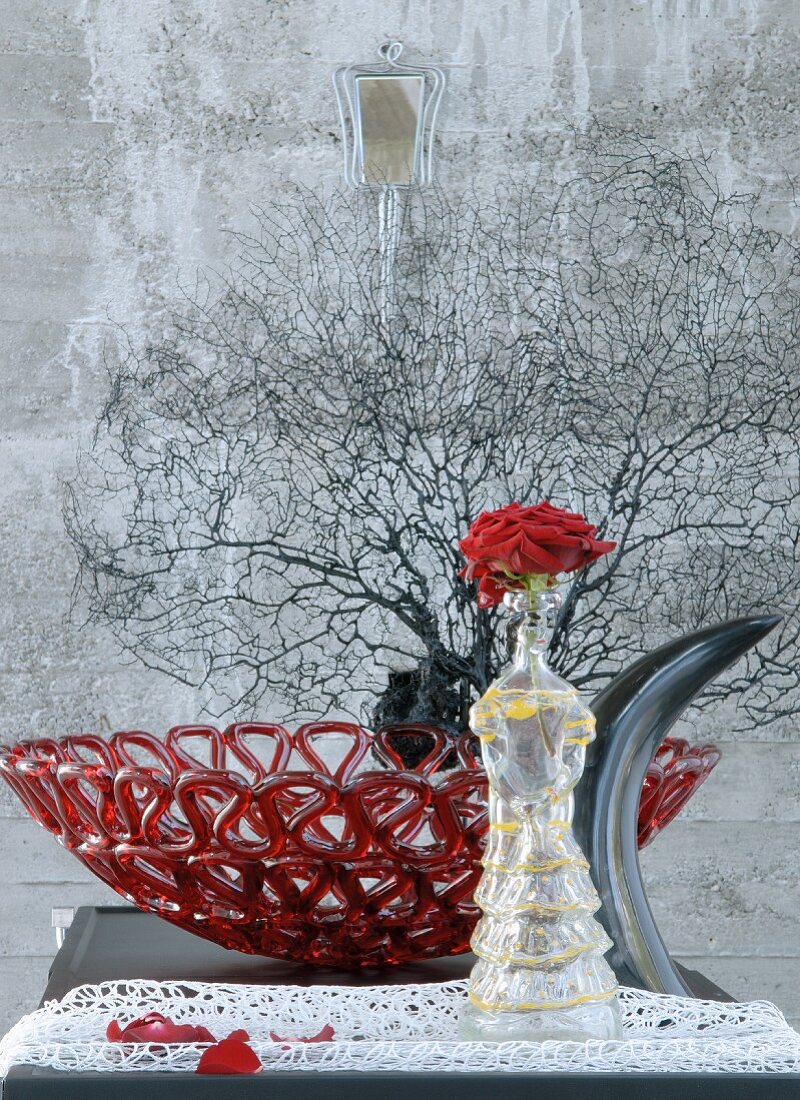 Rose in glass woman-shaped vase in front of red glass dish and black sea fan coral