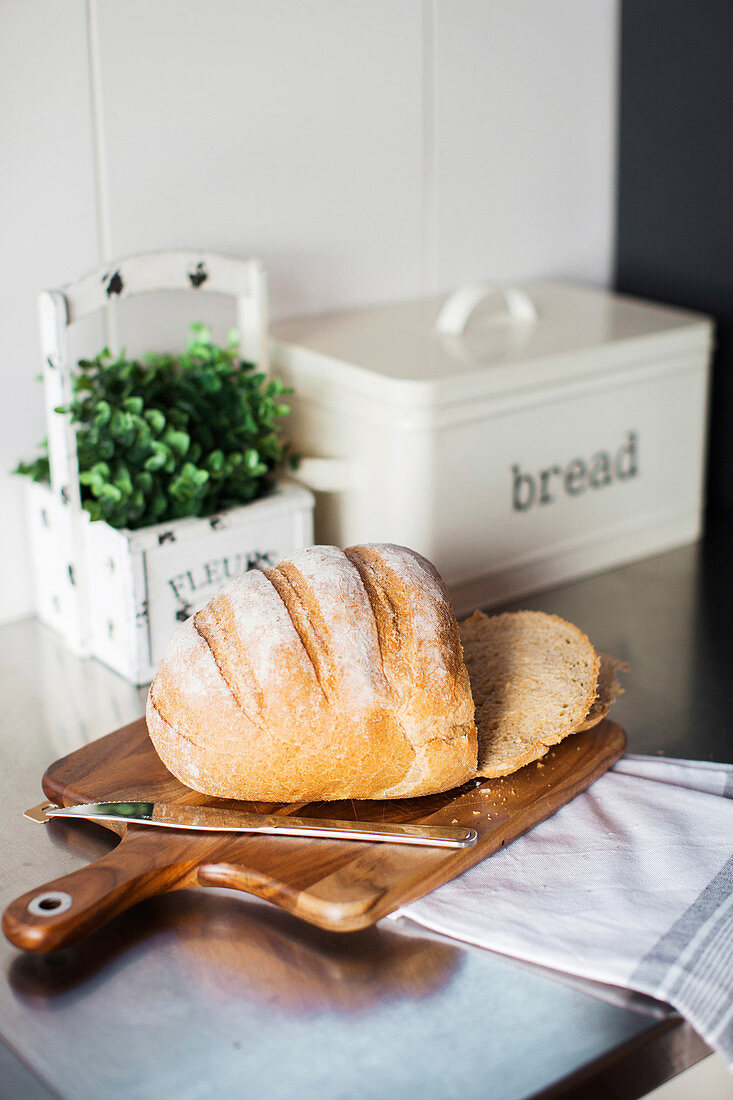 Brot mit Messer auf Holzbrett, im Hintergrund Kräutertopf und Brotkasten