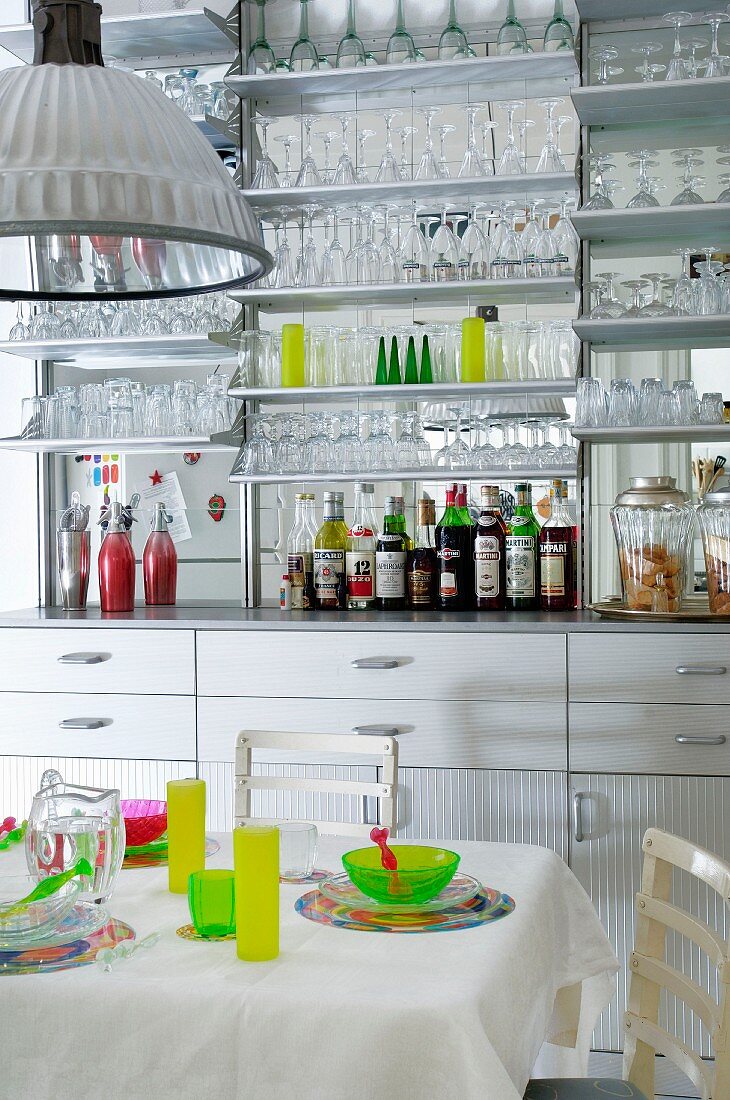 Küchenzeile mit verspiegelter Rückwand und vielen Gläsern auf Wandregal hinter gedecktem Esstisch mit bunten Gedecken
