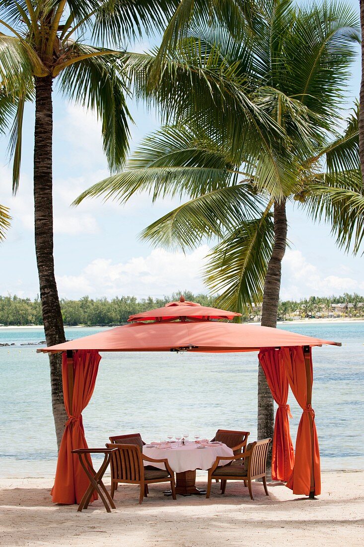 Elegant dining area in orange fabric pavilion under palm trees on beach