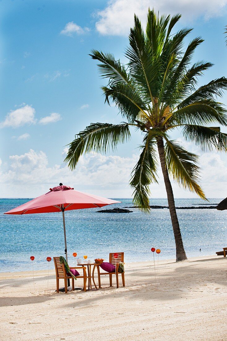 Palm tree, next to table and two chairs below red parasol on idyllic white beach
