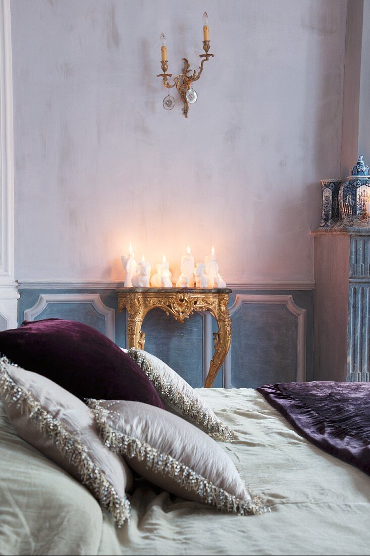 Arrangement of white candles of various shapes on antique gilt console table with scatter cushions on double bed in foreground