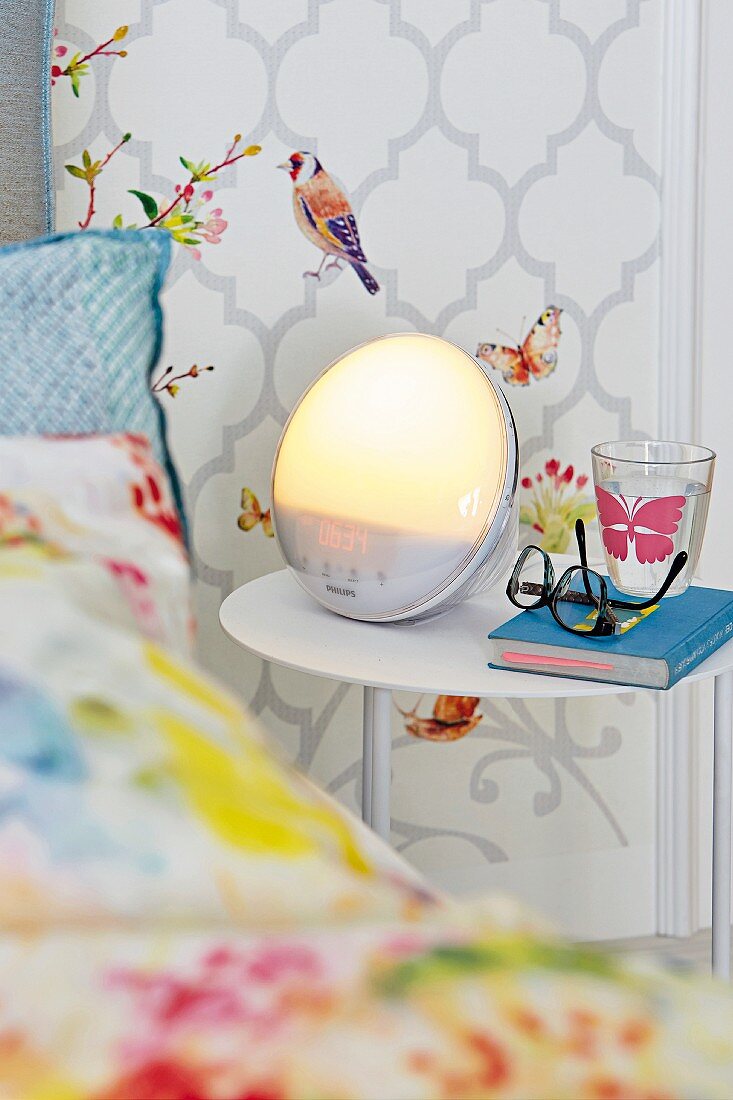 A bedside table with light alarm clock in front of a wall panel with bird wallpaper