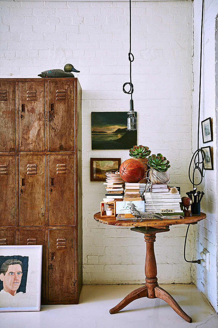 Antique wooden table with books next to vintage metal cupboard in living room with white painted brick wall