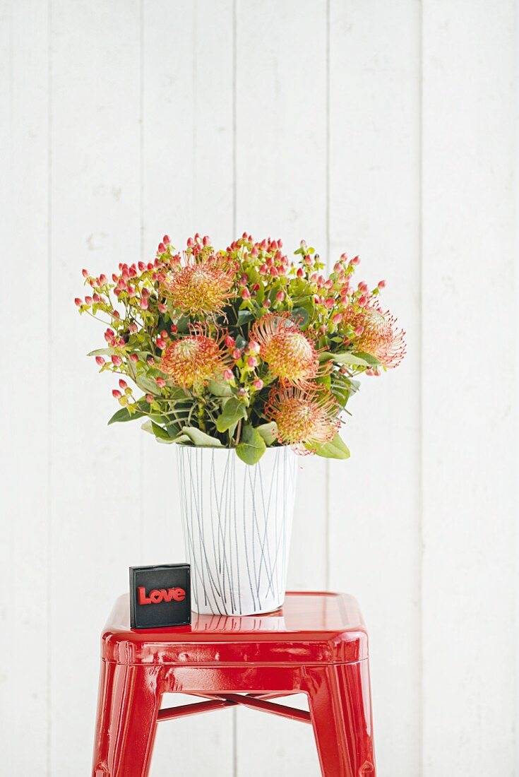Grafisch gestaltetes Papier um Glasvase geklebt und mit Blumenstrauss auf rotem Hocker arrangiert