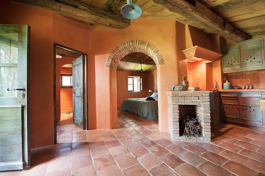 Mediterraner Wohnraum mit Terrakottafliesen und Kamin, Blick durch Rundbogenöffnung ins Schlafzimmer