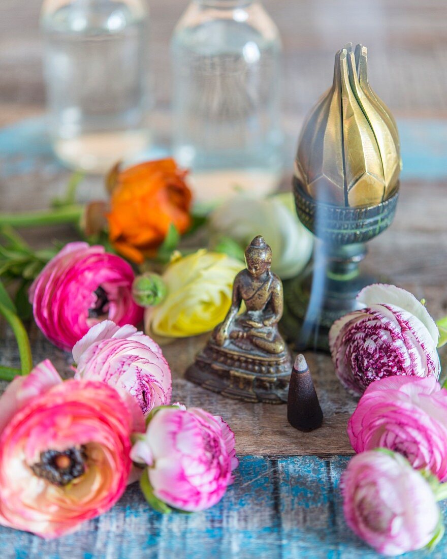 Small Buddha figurine, closed metal incense burner, burning incense and ranunculus flowers on rustic wooden surface