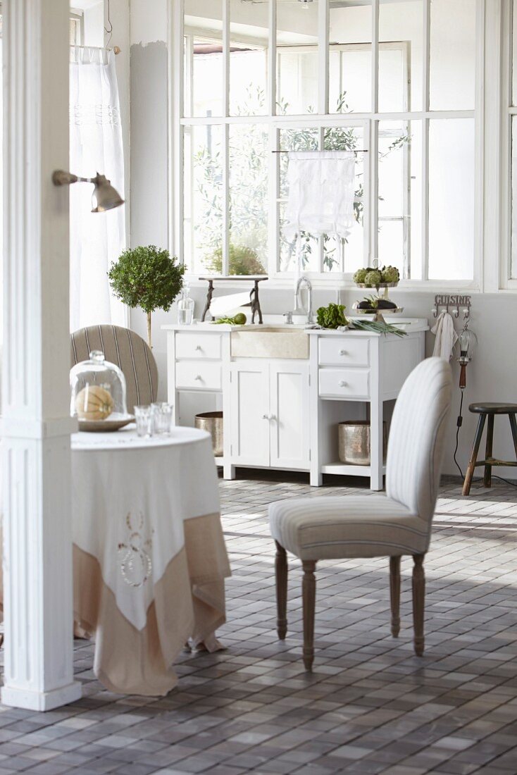 Round table and upholstered chair in front of white kitchen sink and period interior window