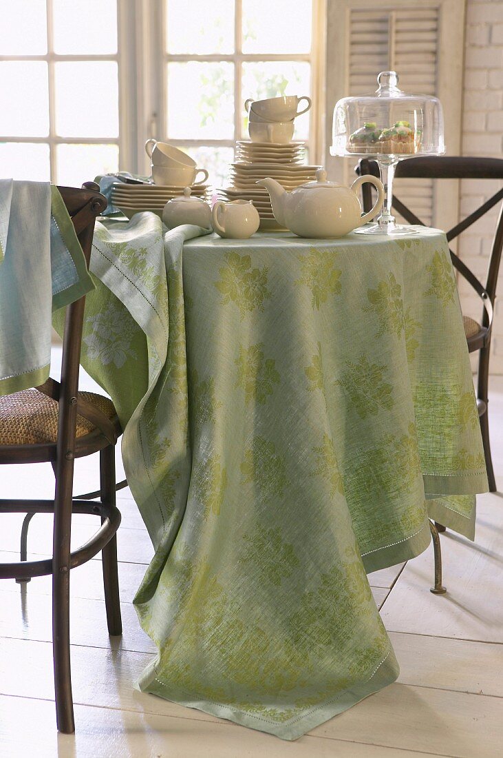 Gestapeltes Porzellan, Teekanne und Glastortenplatte auf rundem Tisch mit pastellgrüner Tischdecke