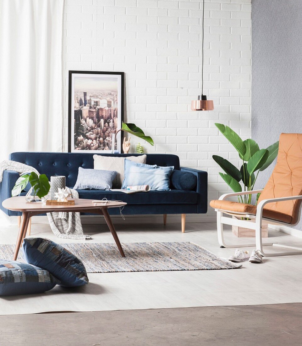 Kissen mit Denim-Bezug auf blauer Couch und auf Boden, Coffeetable, Stuhl mit Leder Auflage, Zimmerpflanze und Pendelleuchte im Wohnzimmer mit Retro Flair