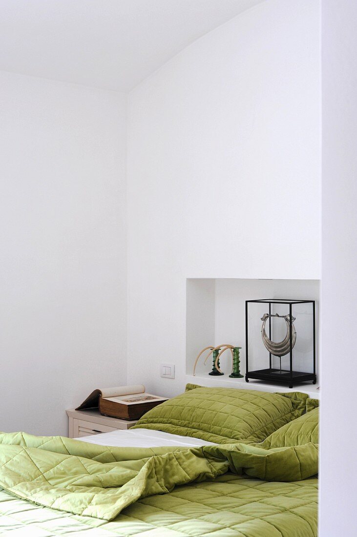 Grüne Bettwäsche neben antiquarischem Buch und Wandnische mit Kunstobjekt am Kopfende
